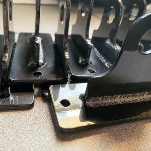 May include: Close-up of several black metal brackets with welded seams. The brackets have a rounded, rectangular shape with a hole in the top corner. The brackets are stacked on top of each other.