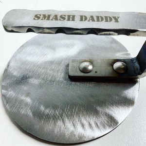 May include: A silver metal burger smasher with a handle. The handle has the words "SMASH DADDY" engraved on it.