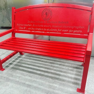 Puede incluir: Un banco de metal rojo con respaldo curvo y una inscripci&oacute;n grabada: "En homenaje a Barbara Bobrich, Profesora de ciencias, Entrenadora de Hawkette, Core&oacute;grafa musical. Miss B... una inspiraci&oacute;n para los estudiantes durante m&aacute;s de 50 a&ntilde;os."