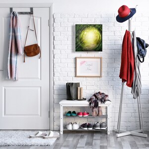 May include: A white brick wall hallway with a white door, a coat rack with a red jacket and a hat, a shoe rack with shoes, and a painting of a green forest path with a light at the end.