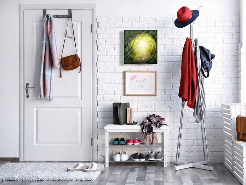 May include: A white brick wall hallway with a white door, a coat rack with a red jacket and a hat, a shoe rack with shoes, and a painting of a green forest path with a light at the end.