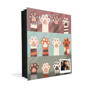 May include: A square glass sign with a black frame featuring a colorful paw print design. The sign has the text "Te quiero, vuelvo pronto" in white lettering. A photo of two people is attached to the sign with magnets.