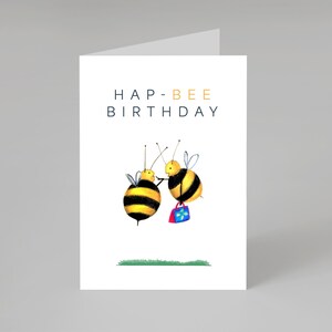May include: A white birthday card with the text "HAP-BEE BIRTHDAY" in navy and gold. The card shows two cartoon bees, one with a shopping bag. A strip of green grass is at the bottom of the card.