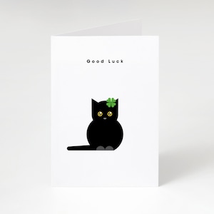 May include: A white greeting card with the words "Good Luck" at the top. The card features a black cat illustration with yellow eyes and a green shamrock on its head. The cat is sitting with its tail extended.