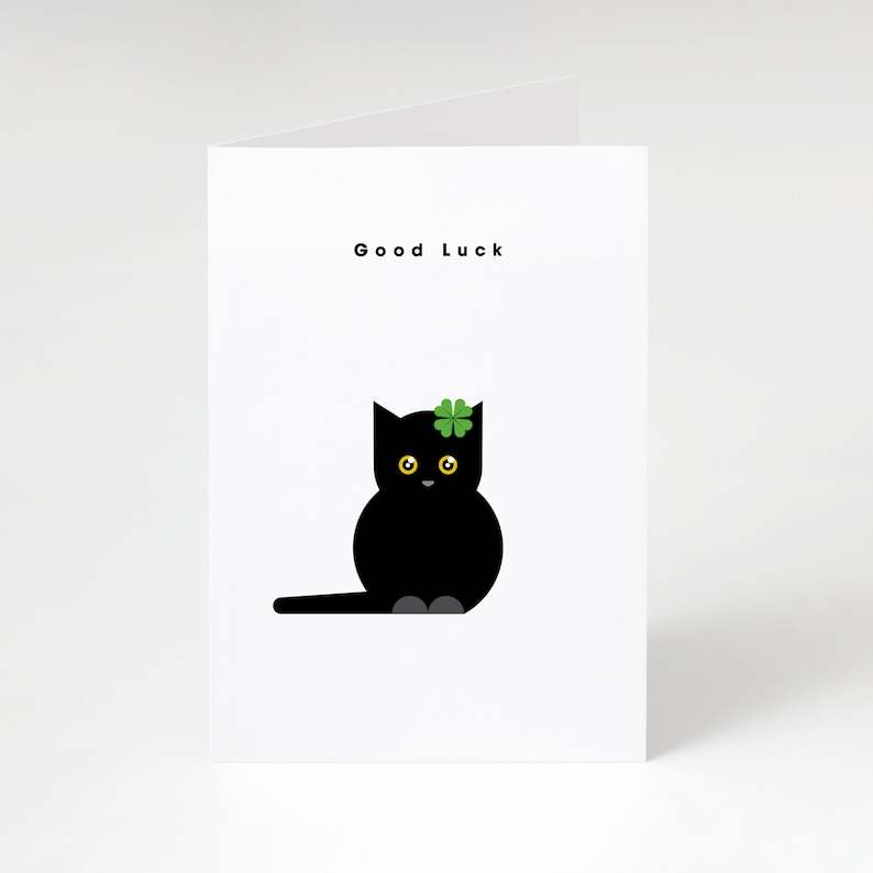 May include: A white greeting card with the words "Good Luck" at the top. The card features a black cat illustration with yellow eyes and a green shamrock on its head. The cat is sitting with its tail extended.