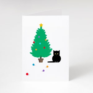 May include: A white Christmas card with a cartoon illustration. It shows a green Christmas tree decorated with baubles and a gold star. A black cat with yellow eyes is next to the tree. Scattered yarn balls.