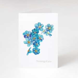 May include: A white card with a watercolour illustration of blue forget-me-not flowers with yellow centres. The words "Thinking of you" are printed at the bottom. The card is blank inside, ready for a personal message.