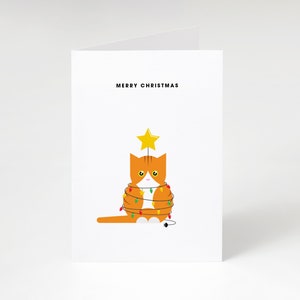 May include: A white Christmas card with the text "MERRY CHRISTMAS". The illustration shows an orange cat decorated as a Christmas tree with fairy lights and a star on top. The cat is the main subject of the card.