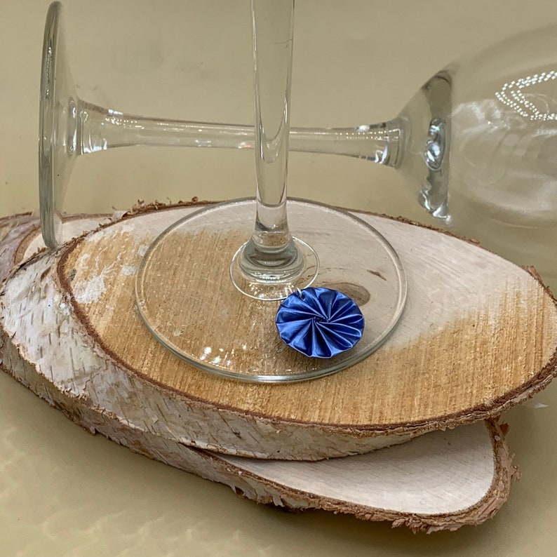 May include: Two clear glass wine glasses on a set of two wooden coasters. The glass on the top has a blue fabric flower charm attached to the stem.