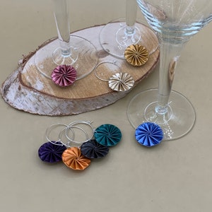 May include: Six colorful wine glass charms with a swirl design. The charms are made of paper and are attached to silver rings. The charms are in the colors blue, green, gold, purple, and pink.