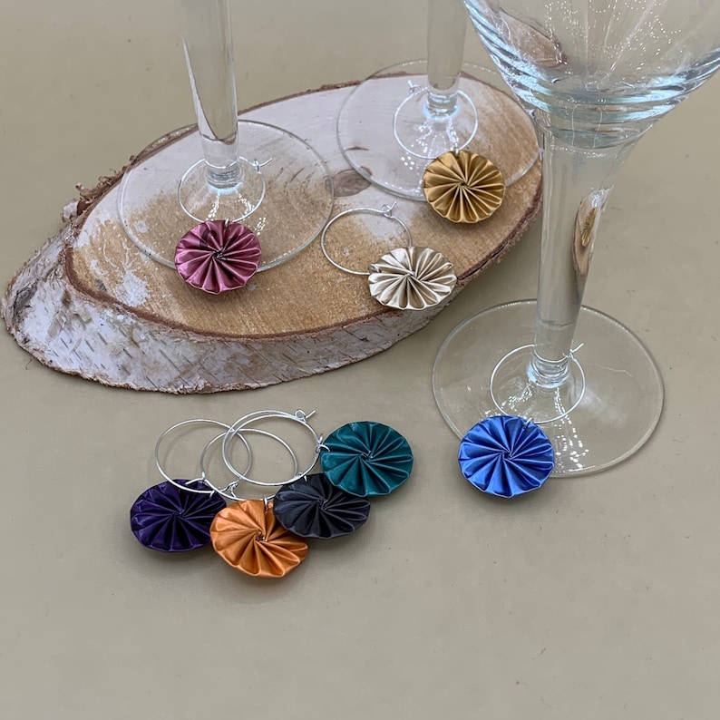 May include: Six colorful wine glass charms with a swirl design. The charms are made of paper and are attached to silver rings. The charms are in the colors blue, green, gold, purple, and pink.