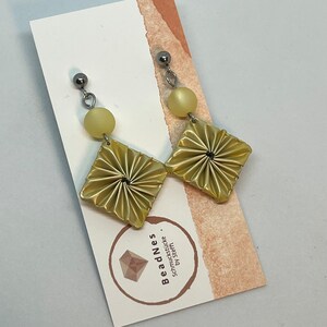 May include: A pair of gold-colored earrings with a pleated fabric design and a small yellow bead on each earring.