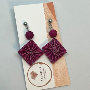May include: A pair of dangle earrings featuring a deep magenta color. Each earring has a small magenta bead and a square, pleated, diamond-shaped pendant. The earrings are attached to a silver-colored post. The earrings are displayed on a white card with text.