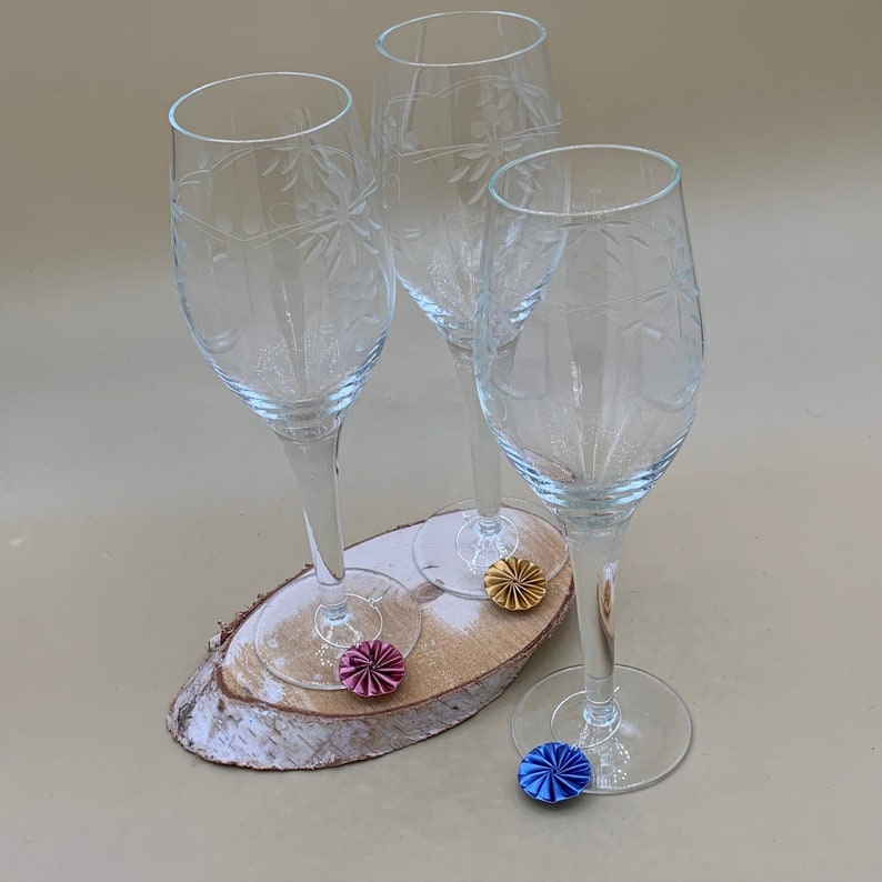 May include: Three clear glass wine glasses with etched designs are arranged on a wooden surface. Each glass has a colorful paper flower attached to the stem.