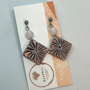 May include: A pair of silver earrings with a white bead and a rose gold square-shaped pendant. The pendant has a pleated design.