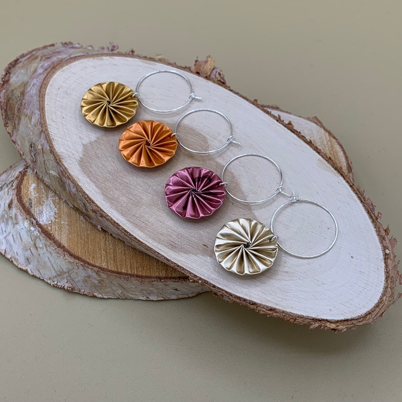 May include: Four pairs of silver hoop earrings with colorful, circular, metallic, paper-like embellishments. The embellishments are in shades of gold, orange, pink, and cream.