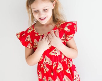 Könnte beinhalten: Ein rotes Kleid mit einem Pizzaschnittenmuster. Das Kleid hat kurze, gerüschte Ärmel und einen runden Ausschnitt. Das Mädchen lächelt und hat die Hände auf der Brust. Der Hintergrund ist eine schlichte weiße Wand.