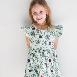May include: A young girl wearing a light green dress with a four-leaf clover and ladybug print. The dress has short, ruffled sleeves and a gathered skirt.