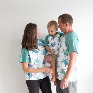 May include: A family wearing matching white t-shirts with a teal green and blue sea turtle print. The father is holding a young boy in his arms. The boy is wearing a teal green and blue sea turtle print t-shirt. The mother is wearing a teal green and blue sea turtle print t-shirt. The family is standing in front of a white wall.
