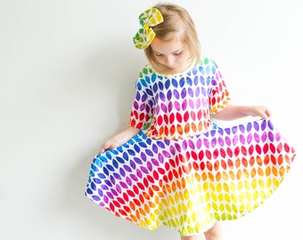 Könnte beinhalten: Ein junges Mädchen trägt ein regenbogenfarbenes Kleid mit weißem Hintergrund und einer weißen Schleife im Haar.