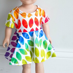 May include: A white dress with a rainbow leaf pattern for a doll. The dress has short sleeves and a flared skirt.