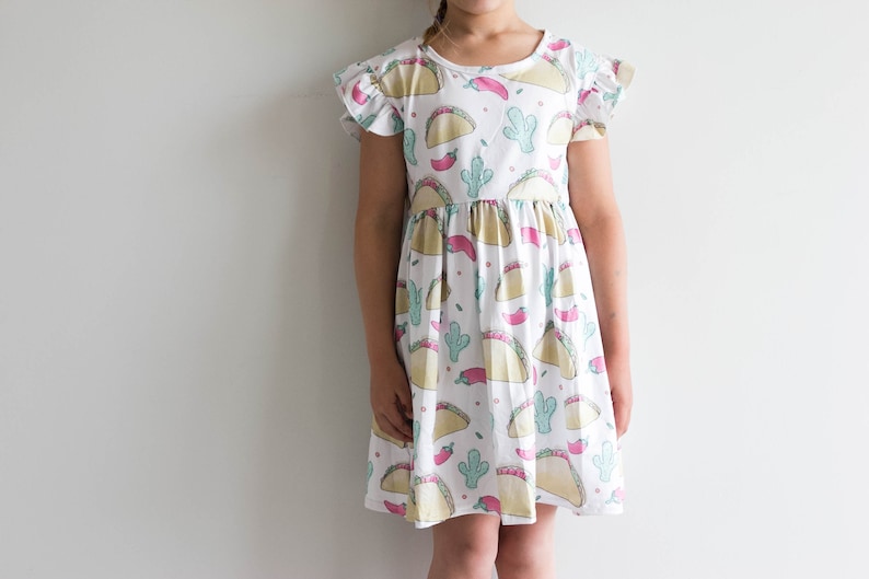 May include: A white dress with a fun print of tacos, cacti, and red peppers. The dress has short sleeves with ruffles and a gathered skirt.