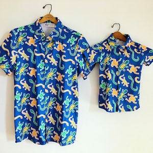 May include: Two blue short-sleeved shirts with a pattern of green and orange lizards on a white background. The shirts are hanging on hangers.