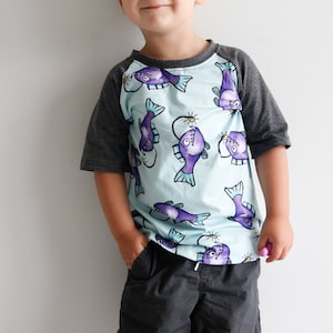 May include: A light blue short-sleeved t-shirt with a black and grey raglan sleeve design. The shirt features a repeating pattern of cartoon anglerfish with purple bodies and yellow lights.