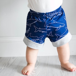May include: A toddler wearing blue and gray shorts with a white shark print. The shorts have a gray elastic waistband and a gray trim around the bottom.