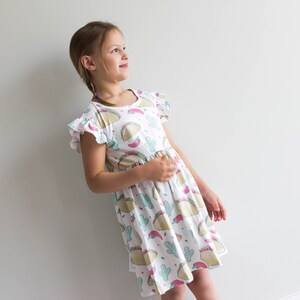 May include: A white dress with a colorful print of tacos, cacti, and chili peppers. The dress has short sleeves with ruffles and a gathered skirt.