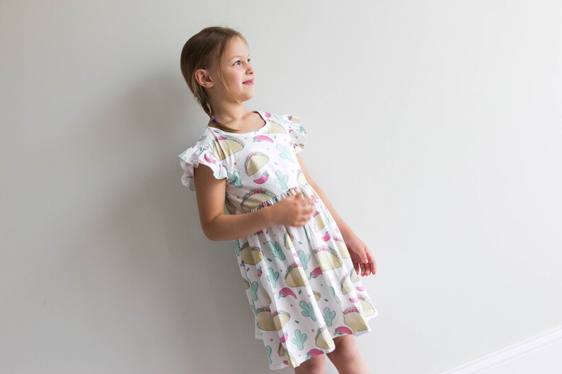 May include: A white dress with a colorful print of tacos, cacti, and chili peppers. The dress has short sleeves with ruffles and a gathered skirt.
