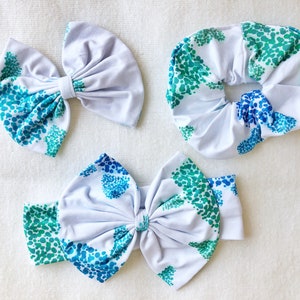 May include: Three white fabric headbands with a blue and green abstract pattern. The headbands are in the shape of a bow, a scrunchie, and a headband with a bow.