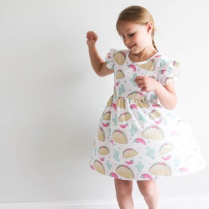 May include: A young girl wearing a white dress with a pattern of tacos, cacti, and chili peppers. The dress has short sleeves and a ruffled hem.