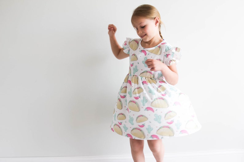 May include: A young girl wearing a white dress with a pattern of tacos, cacti, and chili peppers. The dress has short sleeves and a ruffled hem.