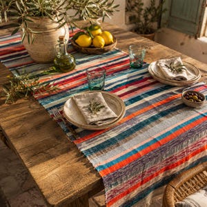 May include: A rustic wooden table is set with a colorful striped table runner. The table is adorned with plates, glasses, and a bowl of lemons. An olive oil bottle and a vase with greenery add to the decor.