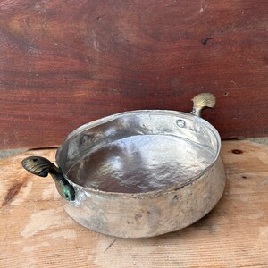 May include: A silver-colored metal bowl with two brass handles. The bowl has a hammered finish and is sitting on a wooden surface.