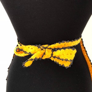 May include: A handmade yellow crochet belt with a black trim and a bow. The belt is tied around a black mannequin. The crochet work is visible, showcasing the texture and craftsmanship of the piece.