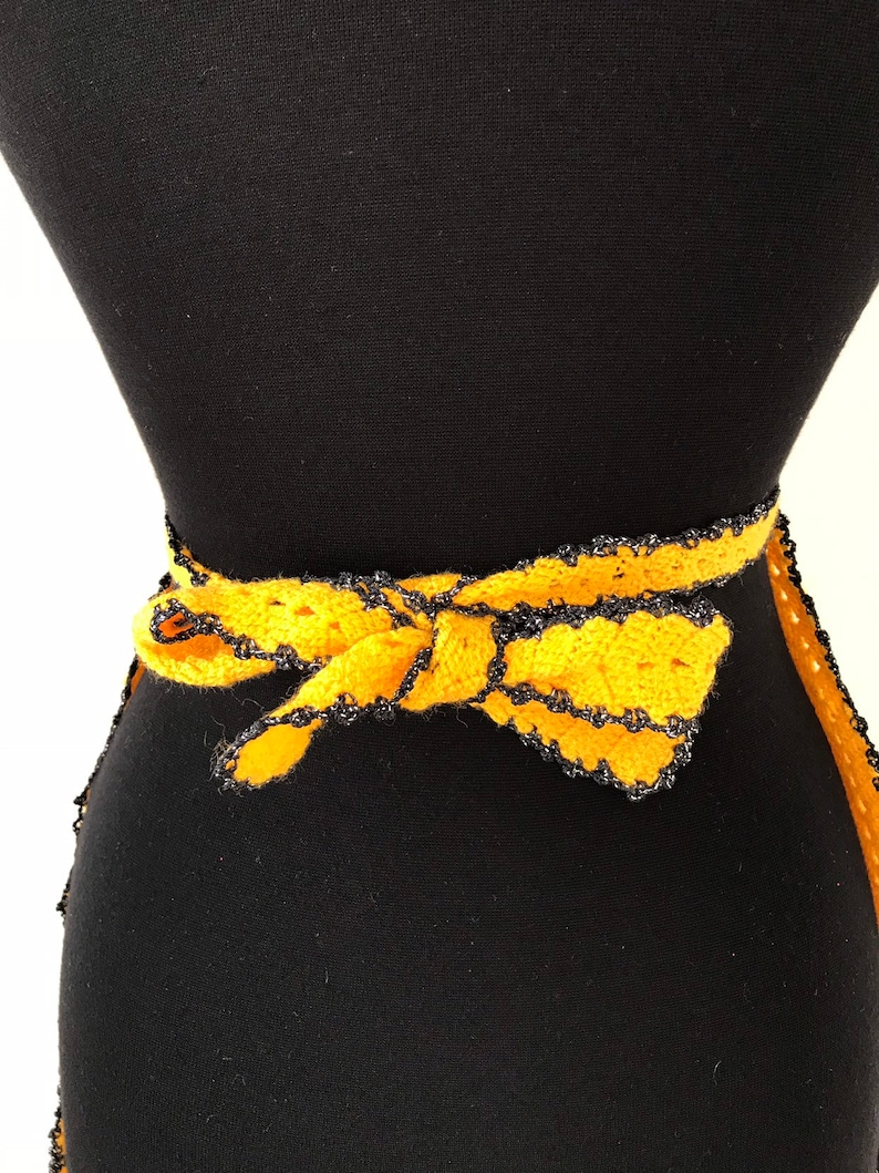 May include: A handmade yellow crochet belt with a black trim and a bow. The belt is tied around a black mannequin. The crochet work is visible, showcasing the texture and craftsmanship of the piece.
