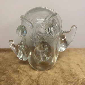 May include: A clear glass sculpture with an abstract, organic shape. The sculpture has a bulbous body with several rounded protrusions and a long, thin spout.