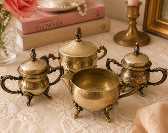 May include: A vintage tea set with a creamer, sugar bowl, and two covered bowls. The set is made of tarnished silver metal with ornate handles and feet. The set is displayed on a white lace tablecloth.