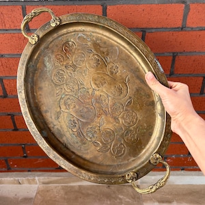 May include: A vintage brass serving tray with ornate floral designs and two handles. The tray is oval shaped and has a slightly worn finish.