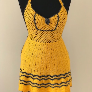 May include: A yellow halter dress with black trim and a decorative black embellishment. The dress is crocheted and features a chevron pattern at the hemline. The dress is displayed on a black mannequin.
