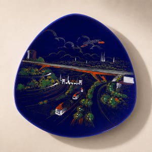 May include: A triangular decorative plate in deep blue featuring a detailed illustration of a cityscape with a prominent bridge, green trees, and buildings. The word LUXEMBOURG is printed in a small banner at the top center of the scene.
