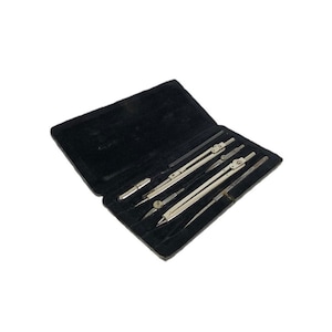 Può includere: Una custodia nera aperta contenente diversi strumenti di disegno argentati. Gli strumenti includono compassi, divisori e uno stilo, tutti ordinatamente disposti contro una fodera di velluto nero. La custodia è rettangolare e sembra vintage.