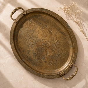 May include: An oval brass serving tray featuring an aged patina finish. The center surface is decorated with an embossed floral pattern, while the rim displays a repeating geometric border. Two ornate, flower-shaped handles are attached to the sides.