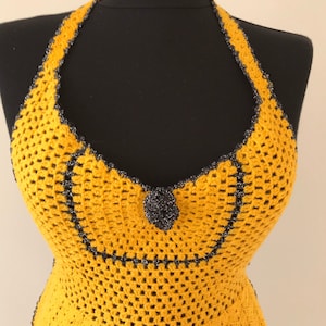 May include: Mustard yellow crochet halter top with a black trim and a decorative black flower. The top is displayed on a black mannequin. The crochet pattern is open and airy.