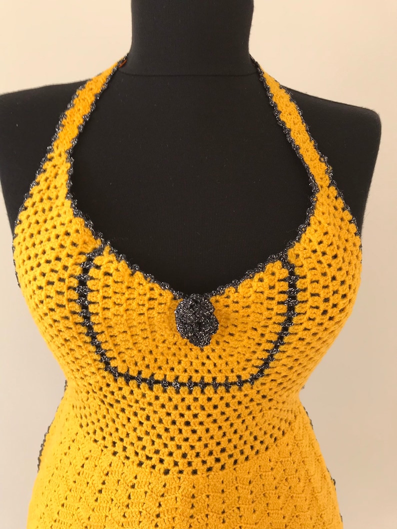 May include: Mustard yellow crochet halter top with a black trim and a decorative black flower. The top is displayed on a black mannequin. The crochet pattern is open and airy.