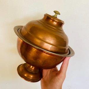 Może przedstawiać: Koperowa misa serwująca z pokrywką i podstawą na nóżkach. Misa ma błyszczące, polerowane wykończenie i prosty, elegancki design.