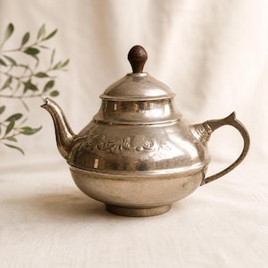 Puede incluir: Una tetera plateada con cuerpo redondeado, boquilla curva y asa. La tapa está rematada con un pomo de madera marrón oscuro. La tetera tiene un diseño floral en relieve alrededor del centro y se apoya sobre una superficie blanca.