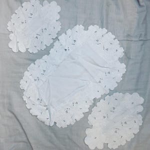 Puede incluir: Un conjunto de tres tapetes blancos bordados con un diseño floral. El conjunto incluye un tapete ovalado grande y dos piezas más pequeñas a juego. Los bordes de cada tapete son festoneados y la tela está ligeramente arrugada.